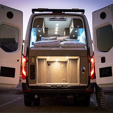 Well-lit camper van interior showing raised bed and illuminated rear gear storage at dusk