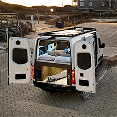 Camper Van parked near beach with open rear doors, pillows on bed, and step laid out on sand