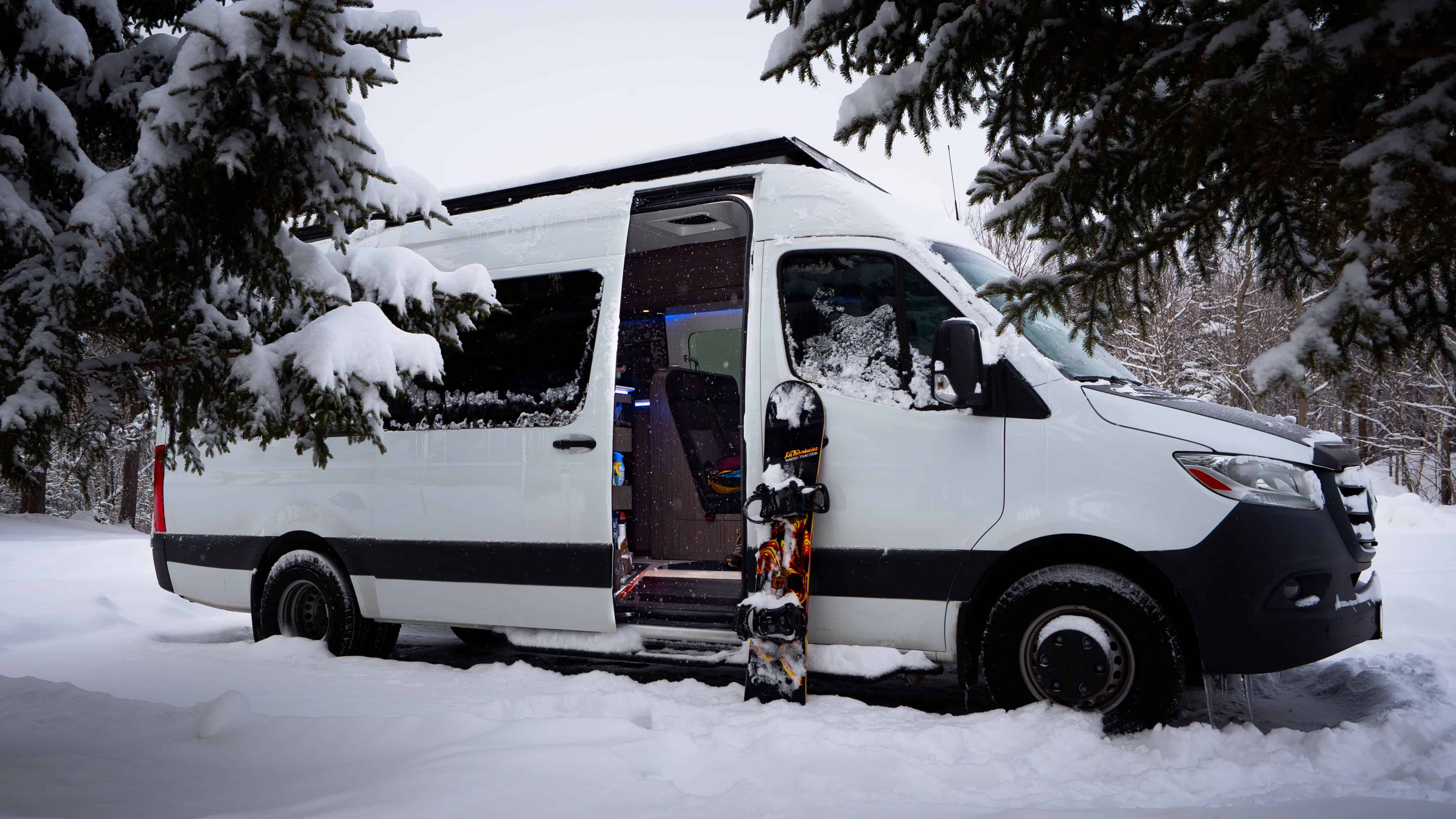 White Icon Vans camper van parked in snowy forest with snowboard by the door